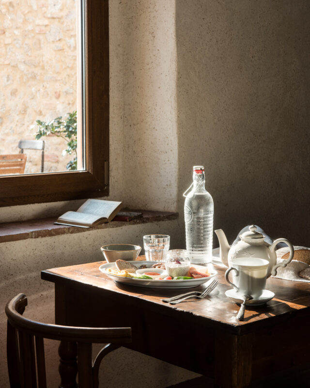A rustic table set with a teapot, glasses, water bottle, and snacks, illuminated by natural light from a nearby window.