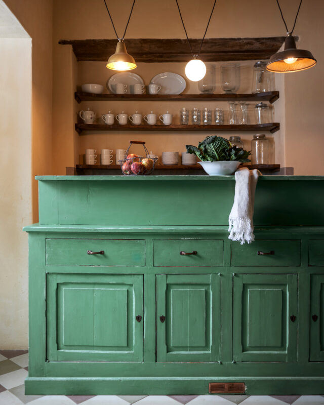 Green kitchen counter with shelves displaying glassware and dishes, illuminated by pendant lights.