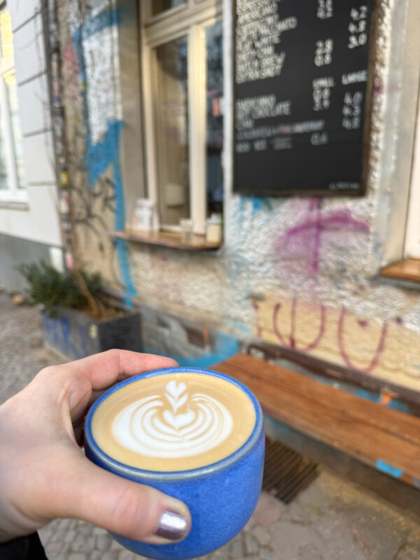 Latte art in a blue cup held in front of a café with a menu board and graffiti on the wall.