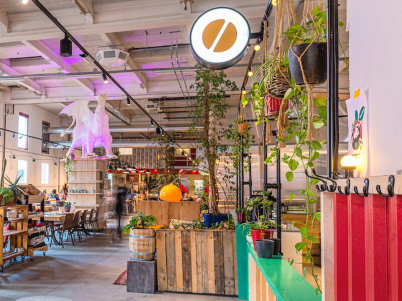 Bright, modern café interior with plants, colorful decor, and patrons seated at wooden tables.