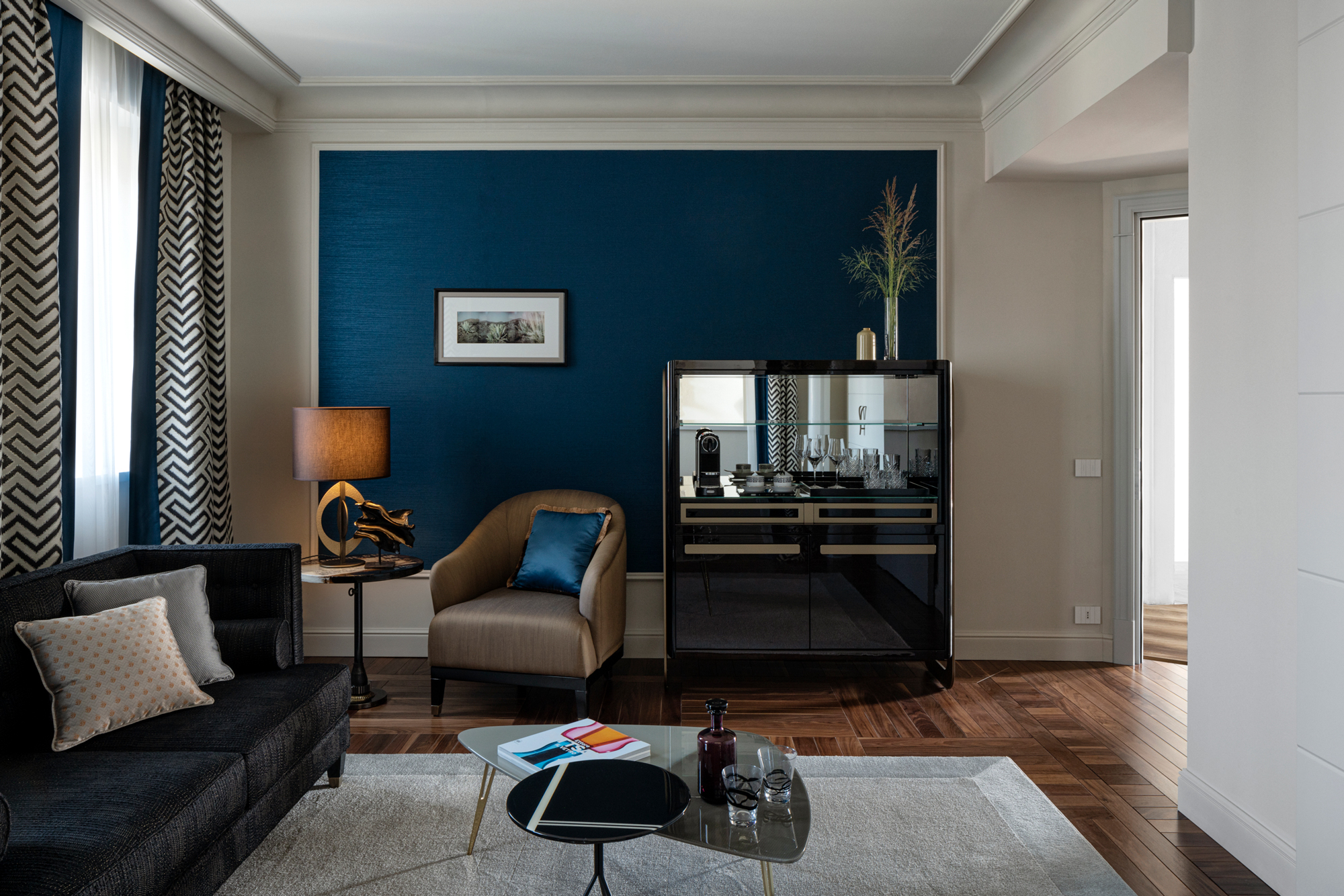 Modern living room featuring a black sofa, beige armchair, blue accent wall, and a glass cabinet with decorative items.