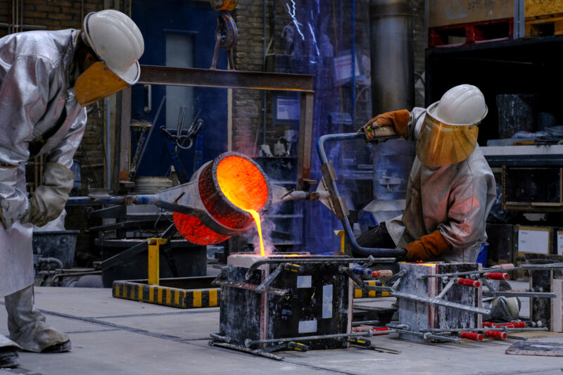 Two workers in protective gear pour molten metal from a crucible into molds in a metalworking shop.