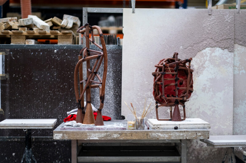 Two abstract sculptures made of twisted materials on a workbench, with tools and a textured wall in the background.
