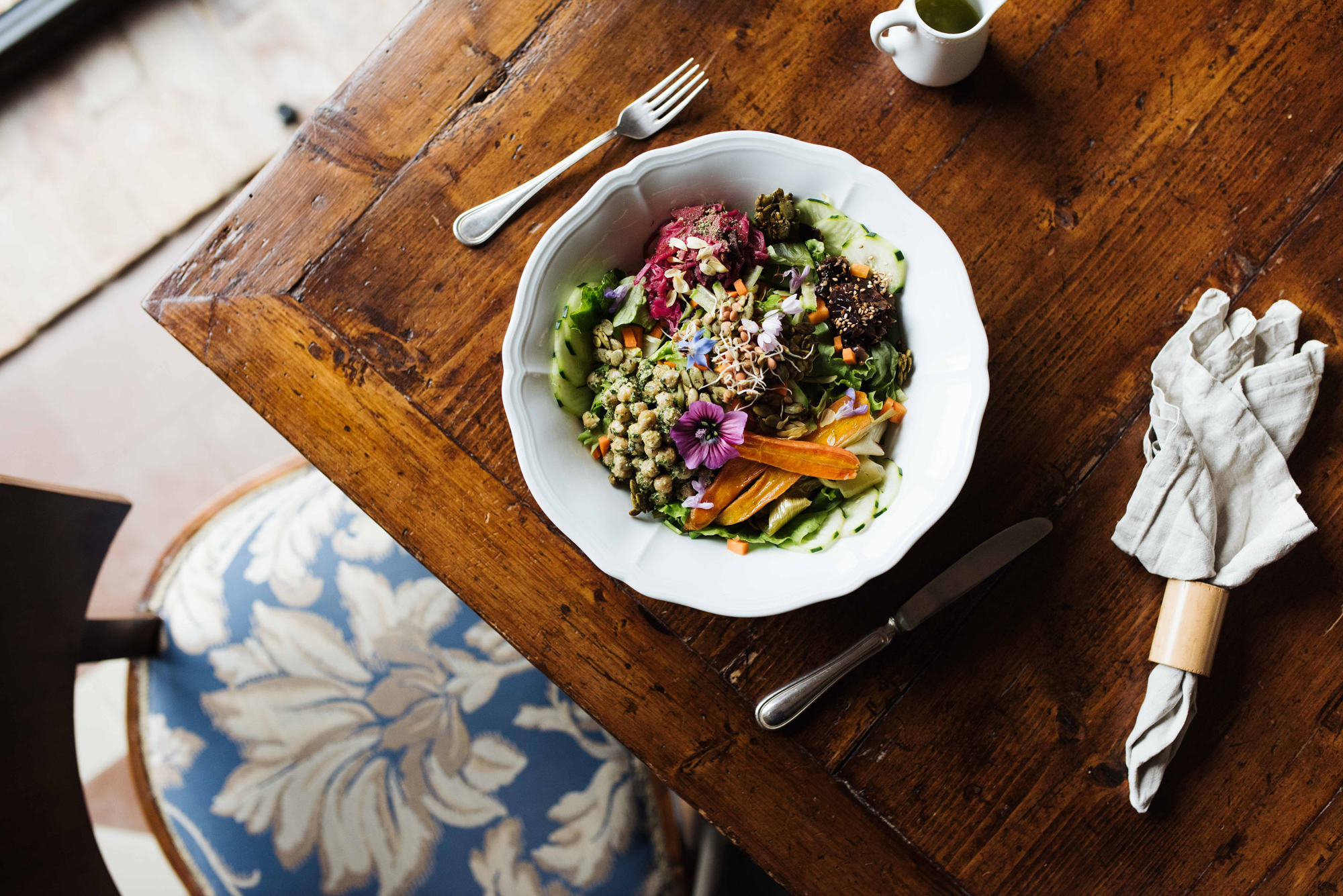 Colorful salad bowl with greens, grains, edible flowers, and vegetables on a wooden table, accompanied by utensils and a s...