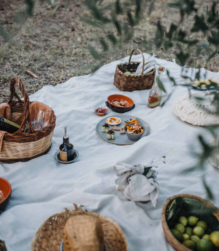 A picnic setup on a white blanket featuring plates of food, baskets, and a bottle, surrounded by greenery.