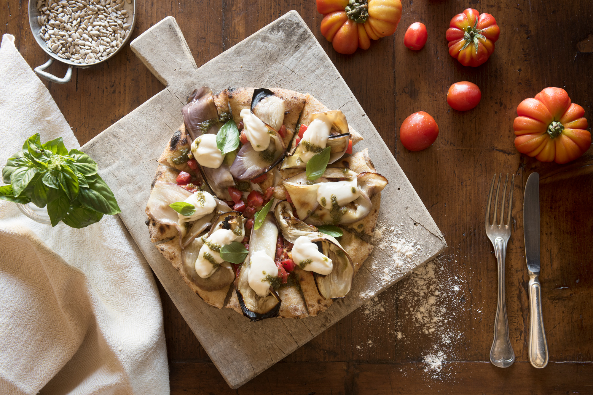 Pizza topped with vegetables, drizzled with sauce, surrounded by tomatoes and basil on a wooden board.