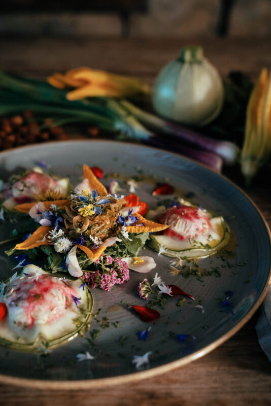 Colorful raw ravioli dish garnished with edible flowers and herbs, surrounded by fresh vegetables on a wooden table.
