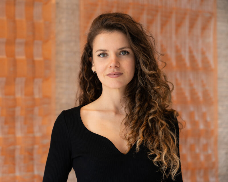 Young woman with long, curly hair, wearing a black top, stands in front of textured orange wall art.