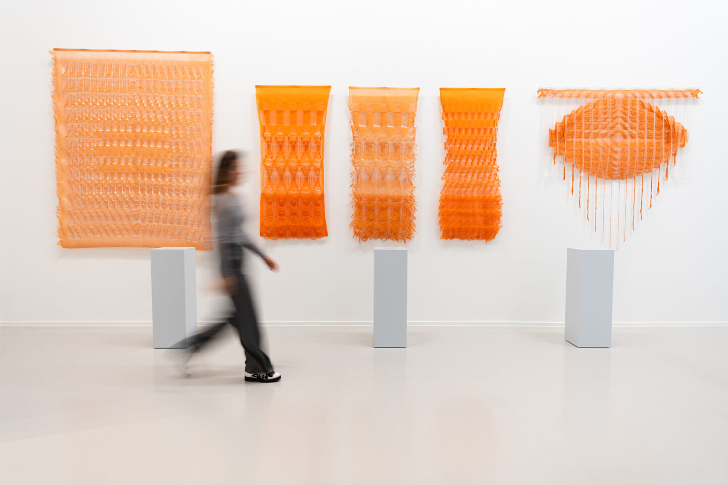 Abstract orange textile artworks displayed on white walls, with a figure walking past.