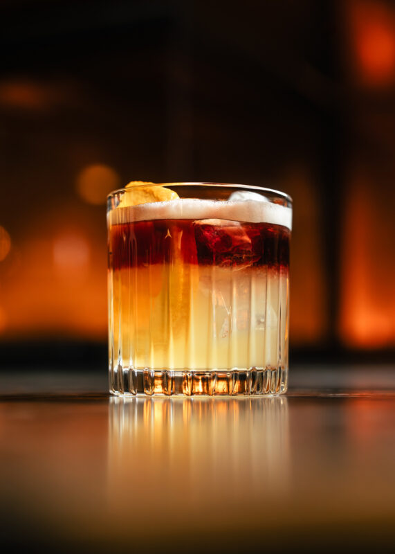 Layered cocktail in a clear glass, featuring dark and light colors with a garnish of citrus peel.