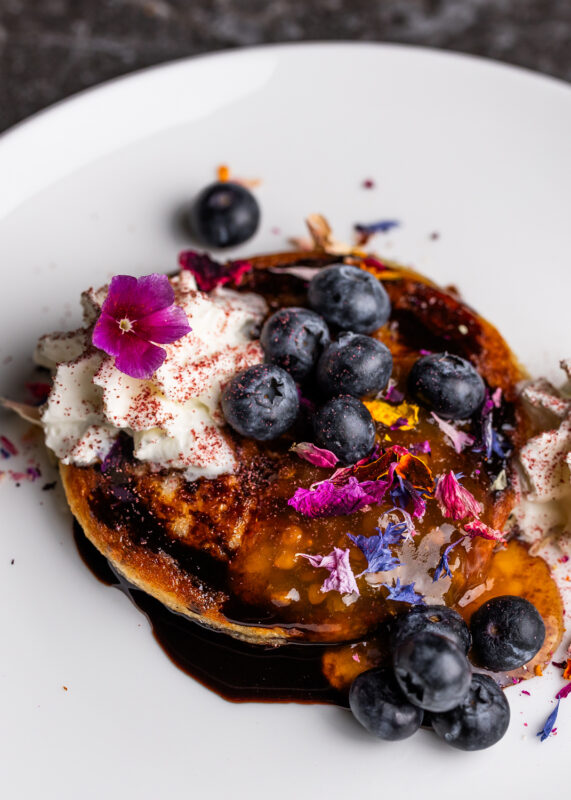 Pancakes topped with blueberries, whipped cream, edible flowers, and a drizzle of syrup on a white plate.