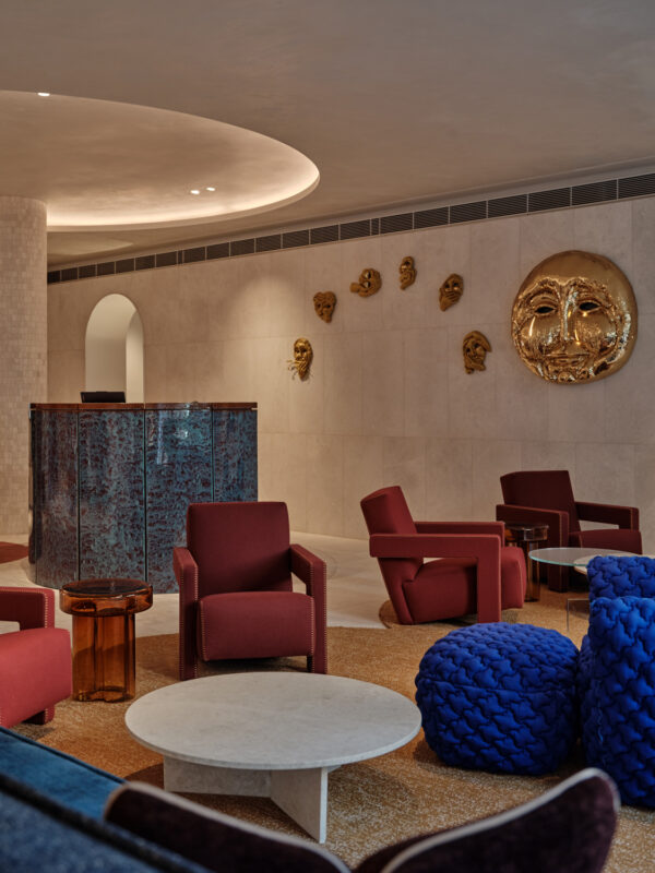 Stylish hotel lobby featuring red chairs, blue poufs, a marble table, and a decorative wall with gold masks.