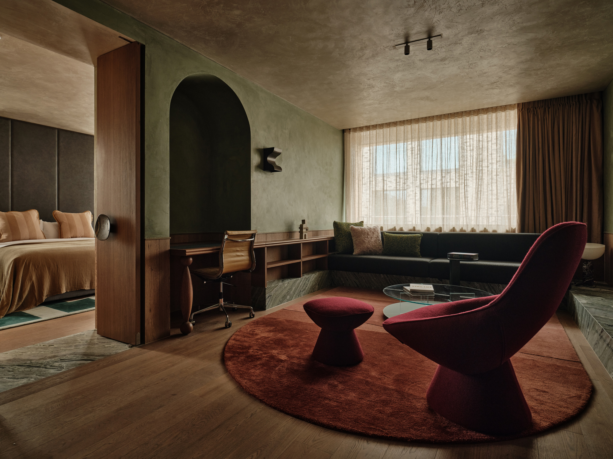 Stylish hotel suite featuring a cozy seating area, desk, and large window with sheer curtains.