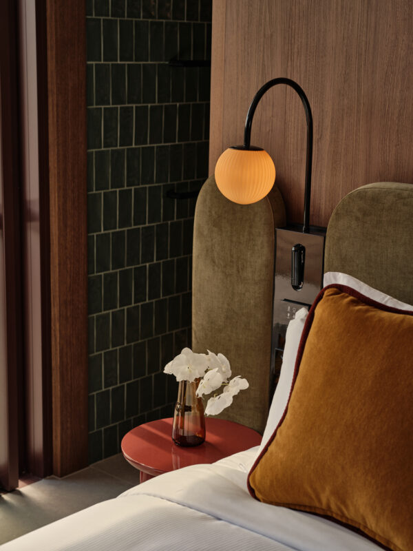 Elegant hotel room featuring a bedside table with a vase of white flowers, a stylish lamp, and a plush orange pillow.