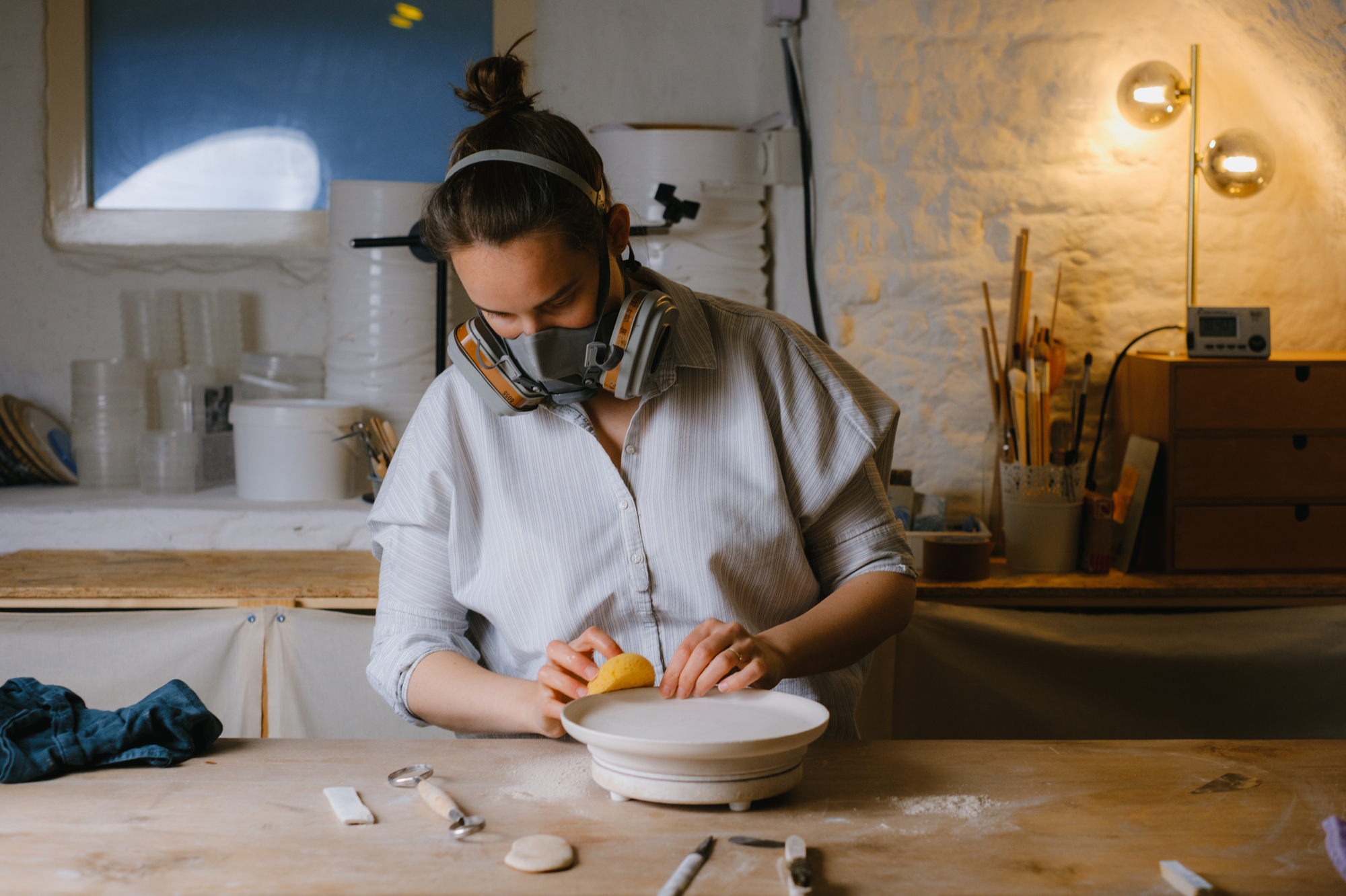 Woman wearing a mask and headphones shapes clay on a pottery wheel in a well-lit studio.
