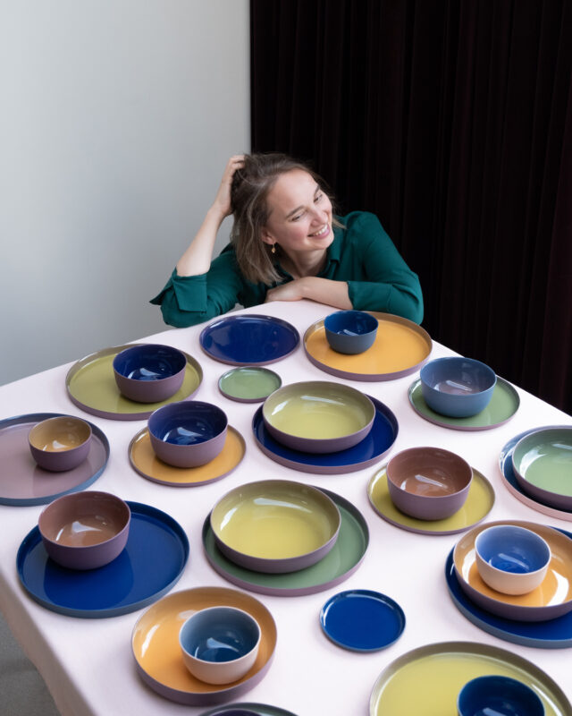 Woman with short hair smiles while sitting at a table adorned with colorful ceramic plates and bowls.