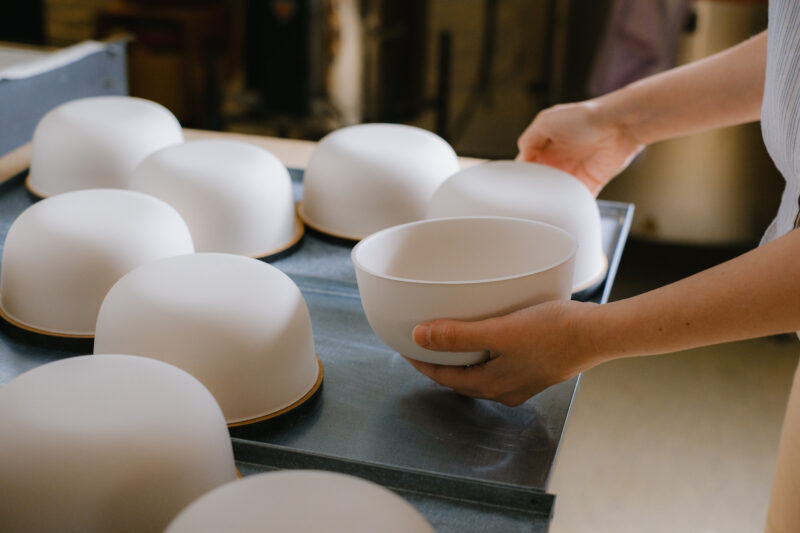 Hands placing a white bowl among several white ceramic bowls on a tray in a studio setting.