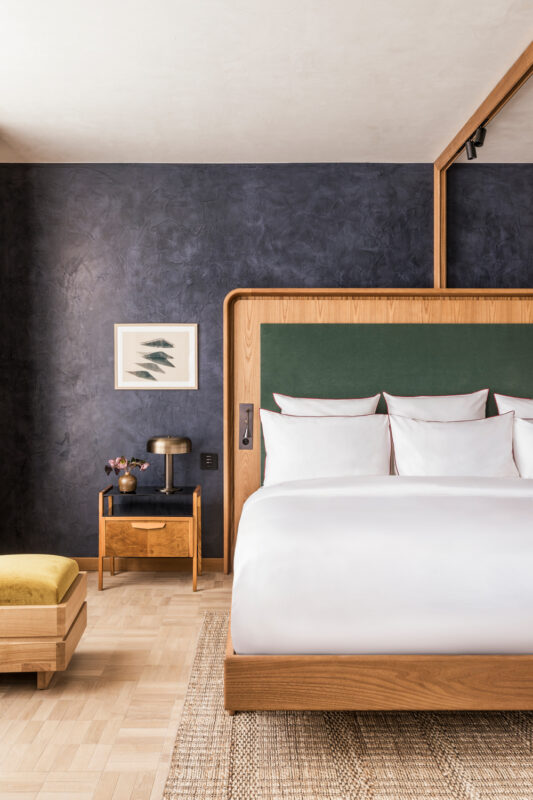 Modern hotel room featuring a wooden bed with green headboard, side table, and textured wall.