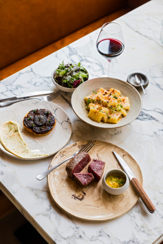 Gourmet meal featuring steak, salad, and sides on a marble table, with a glass of red wine.
