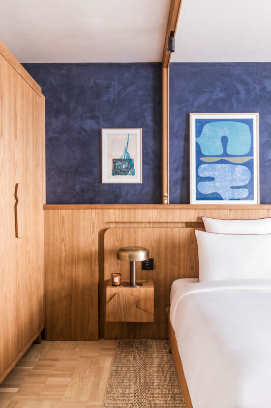 Stylish hotel room featuring wooden furniture, a bedside table with a lamp, and framed artwork on a blue wall.
