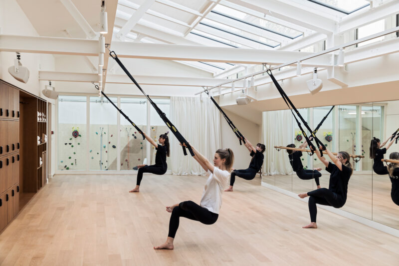 Women practicing suspension training in a bright, modern fitness studio with large windows and wooden floors.