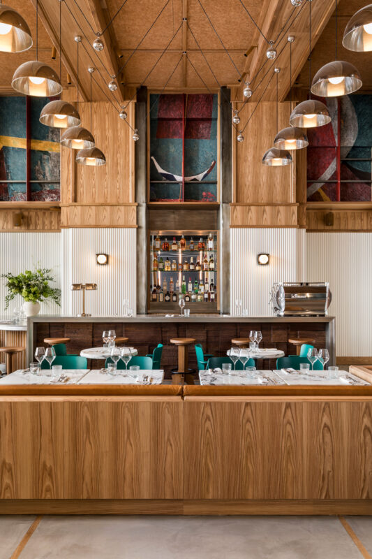 Modern brasserie interior with wooden accents, green chairs, and a bar displaying various bottles. Colorful artwork on the...
