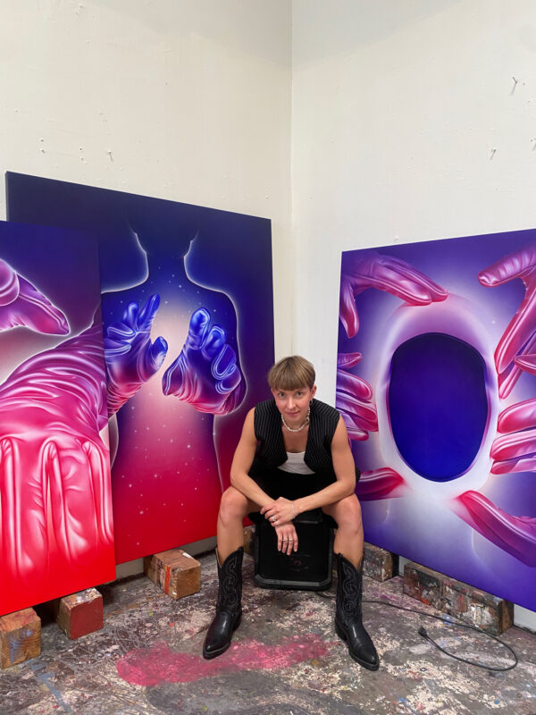 Aistė Stancikaitė sits on a stool in an art studio, surrounded by colorful paintings featuring hands and a circular shape.