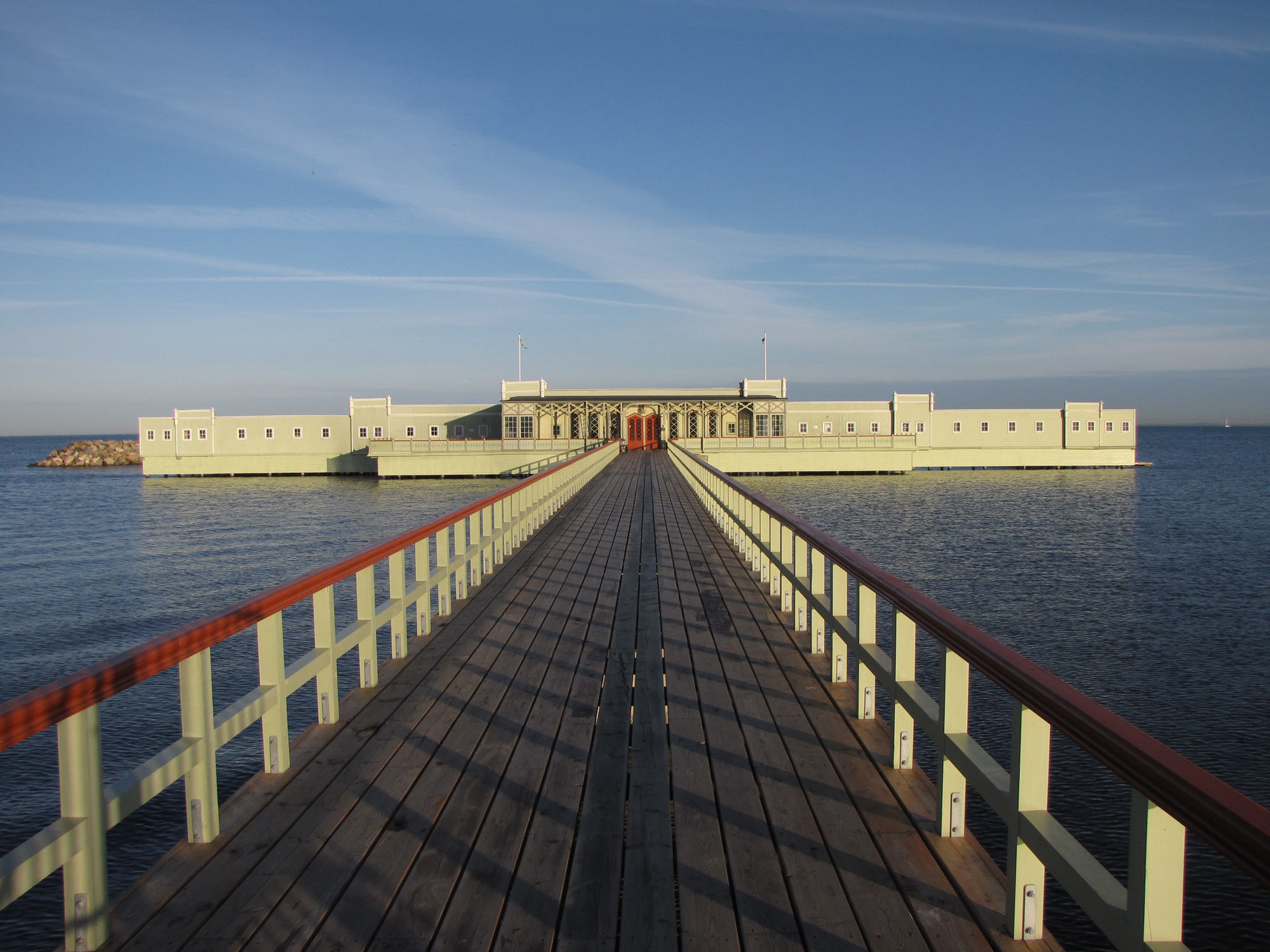 Wooden pier leading to a light green building on water, under a clear blue sky.