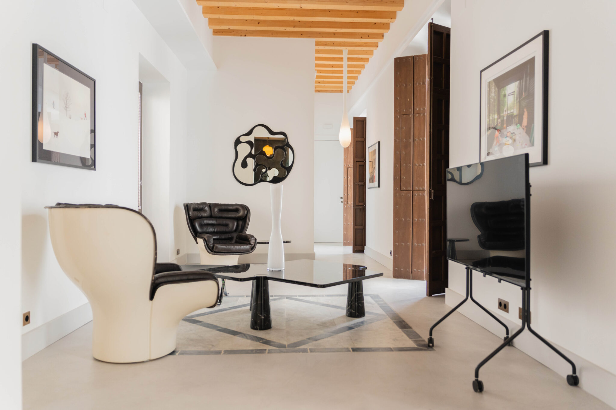 Modern living room featuring a black and white chair, a glass coffee table, and a TV on a stand, with wooden beams above.