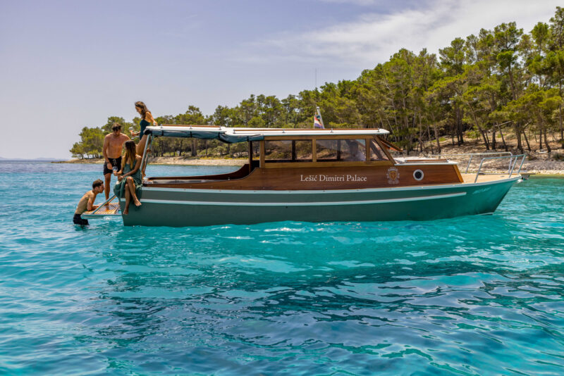 Wooden boat named "Lešić Dimitri Palace" anchored in clear blue water, with people climbing aboard from the side.