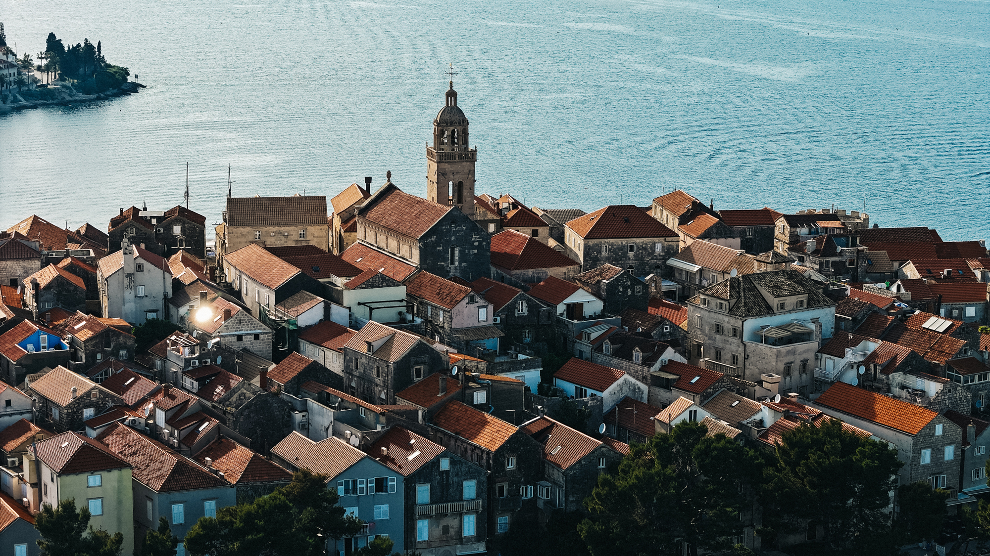 Aerial view of Korčula town, featuring red-roofed buildings and a church, with the sea in the background.