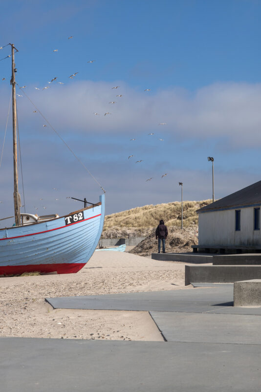 A blue fishing boat on a sandy beach, with a person standing nearby and seagulls flying overhead.