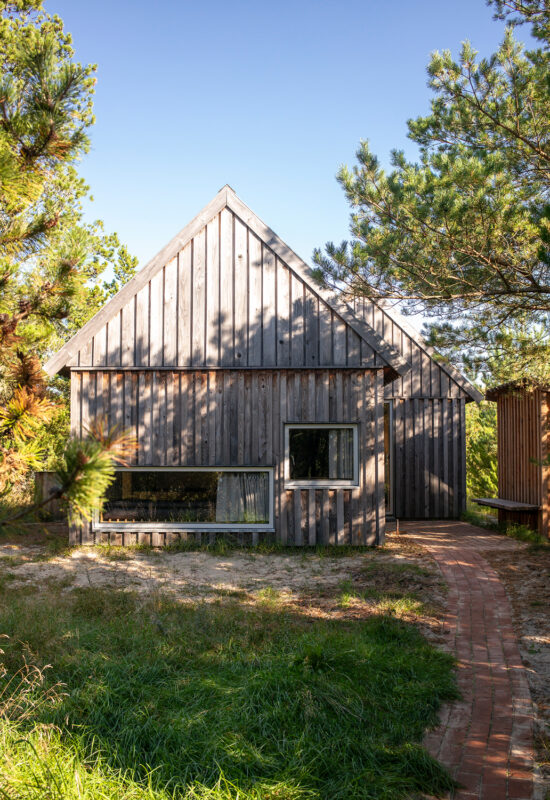 Wooden cabin with a triangular roof, surrounded by greenery and a brick pathway leading to the entrance.