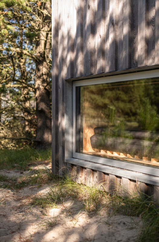Wooden building corner with a large window reflecting trees and grass, set in a sandy area.