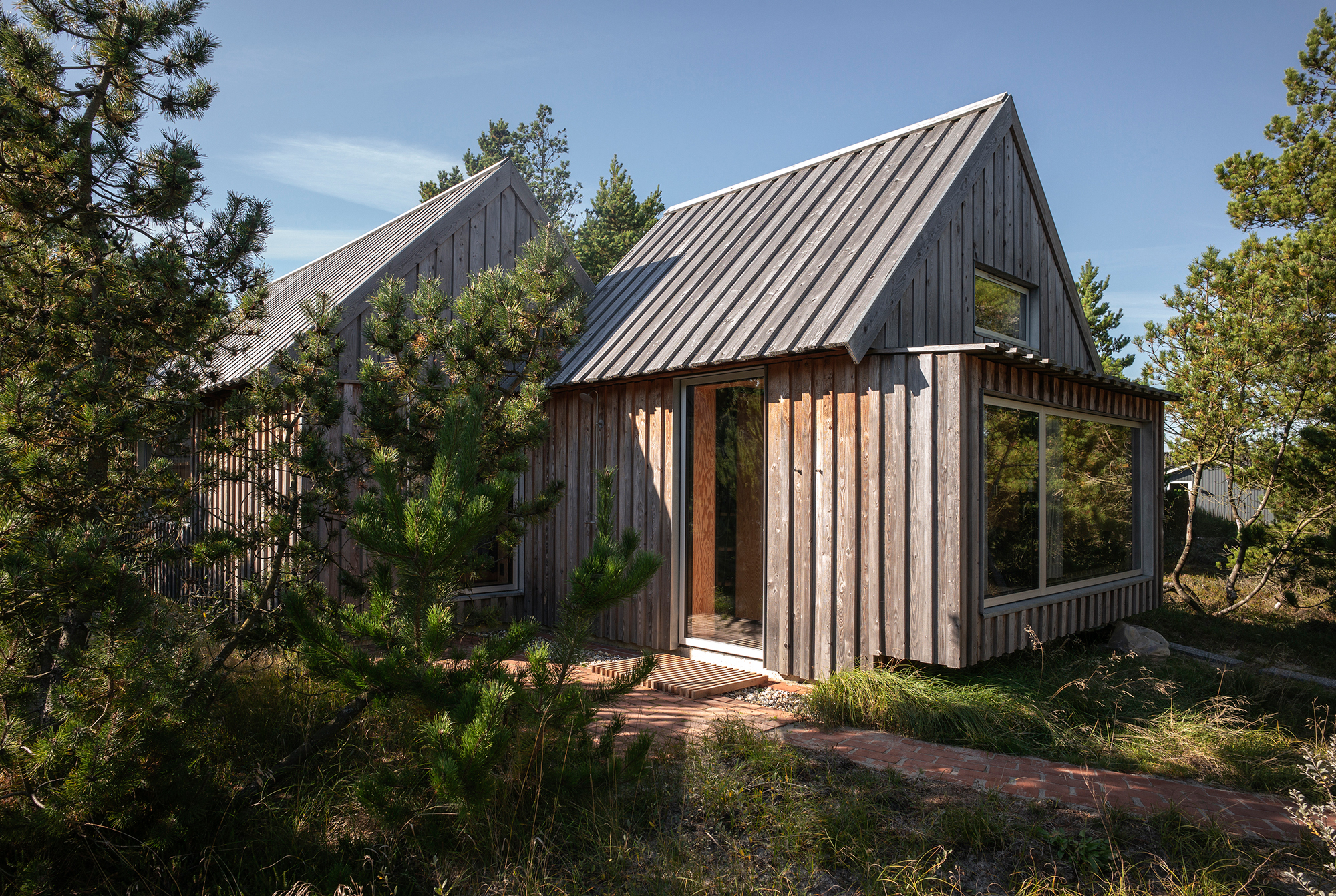 Wooden cabins with sloped roofs surrounded by greenery and a brick pathway, set in a natural landscape.
