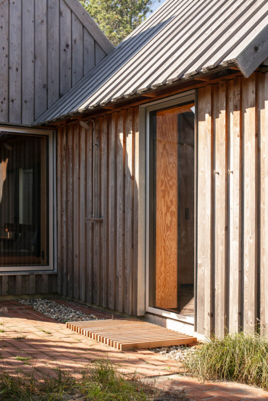 Wooden exterior of a modern building with a sloped roof, featuring a glass door and wooden slats on the ground.