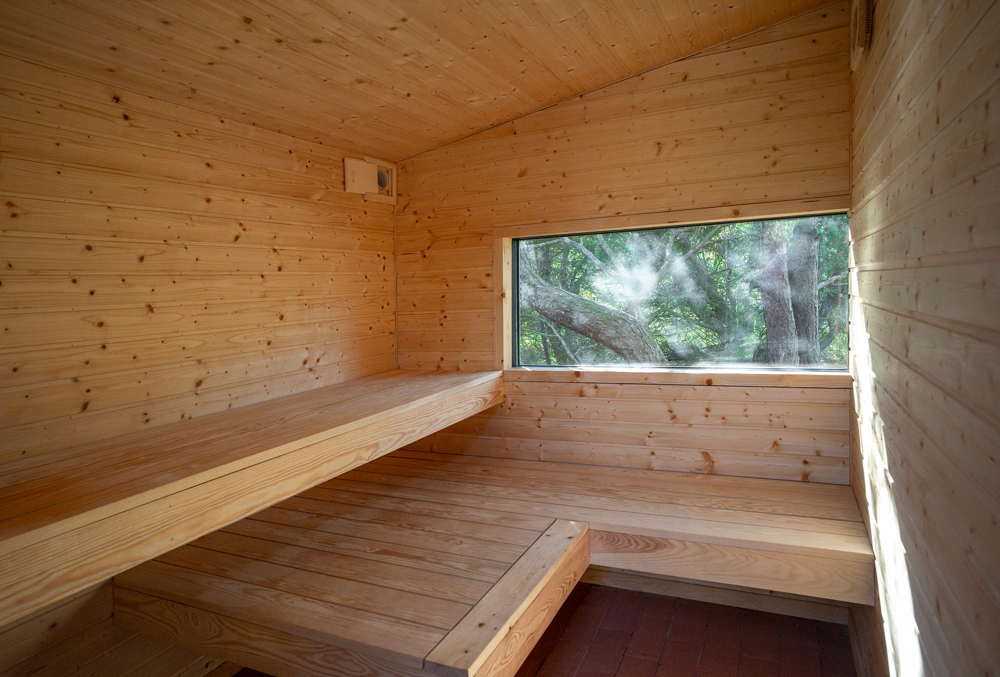 Wooden sauna interior with benches and a large window overlooking trees. Natural light filters through the glass.