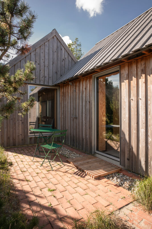 Wooden cabin exterior with a patio featuring a green table and chairs, surrounded by grass and trees under a blue sky.
