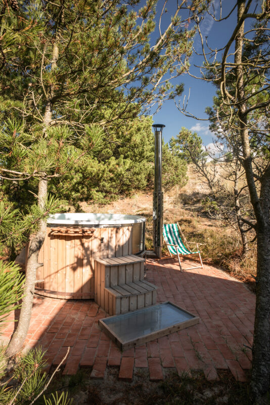 Wooden hot tub with steps, surrounded by trees, next to a striped lounge chair on a brick patio.