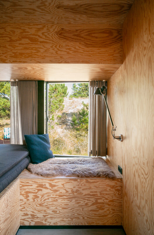 Cozy interior of a wooden cabin with a window view, featuring a bed nook and a reading lamp.
