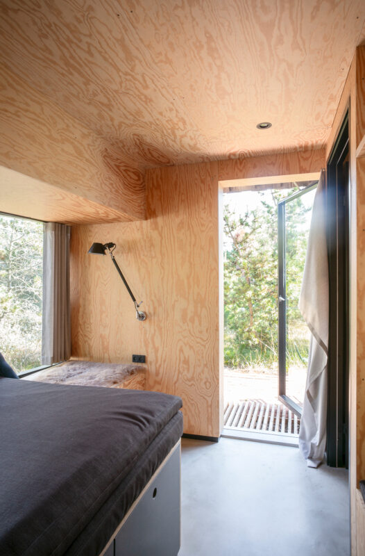 Cozy interior of a wooden cabin with a bed, lamp, and open door leading to a green outdoor area.