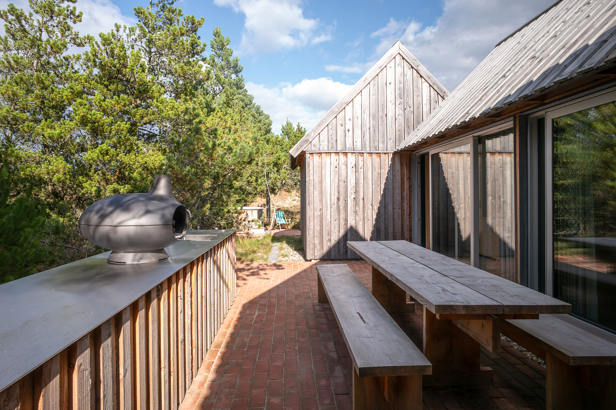 Wooden outdoor seating area with a metal fireplace, surrounded by trees and a modern wooden building.