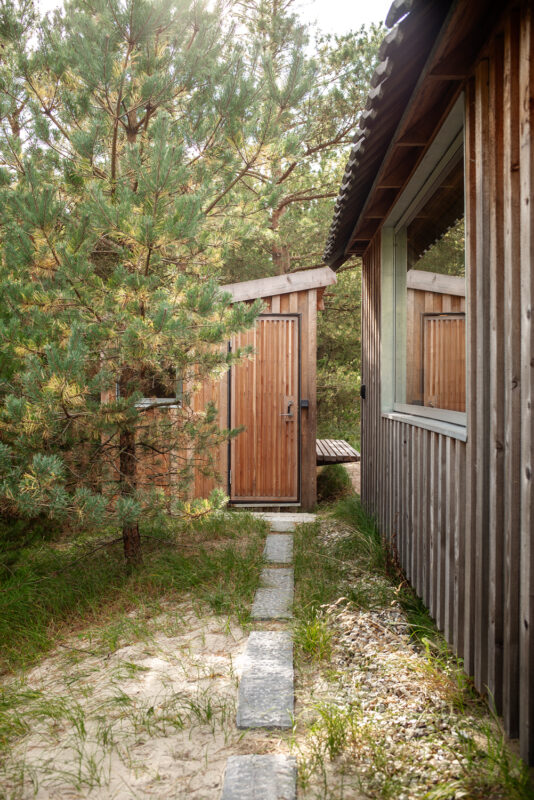 Wooden pathway leading to a door surrounded by trees, with a cabin visible on the right.
