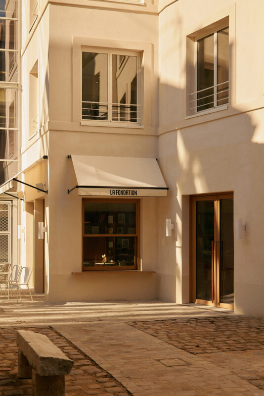 Modern building exterior with a window labeled "LA FONDATION," featuring a sunlit courtyard and stone seating.