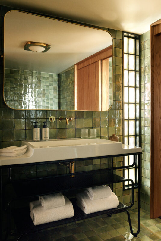 Modern bathroom featuring green tiled walls, a large mirror, a white sink, and neatly stacked towels on a black metal shelf.