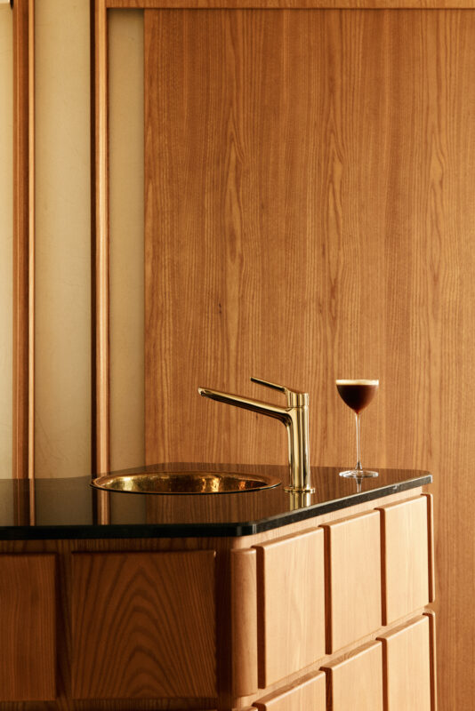 Elegant bar area featuring a gold faucet, black countertop, and a glass of dark beverage against a wooden backdrop.