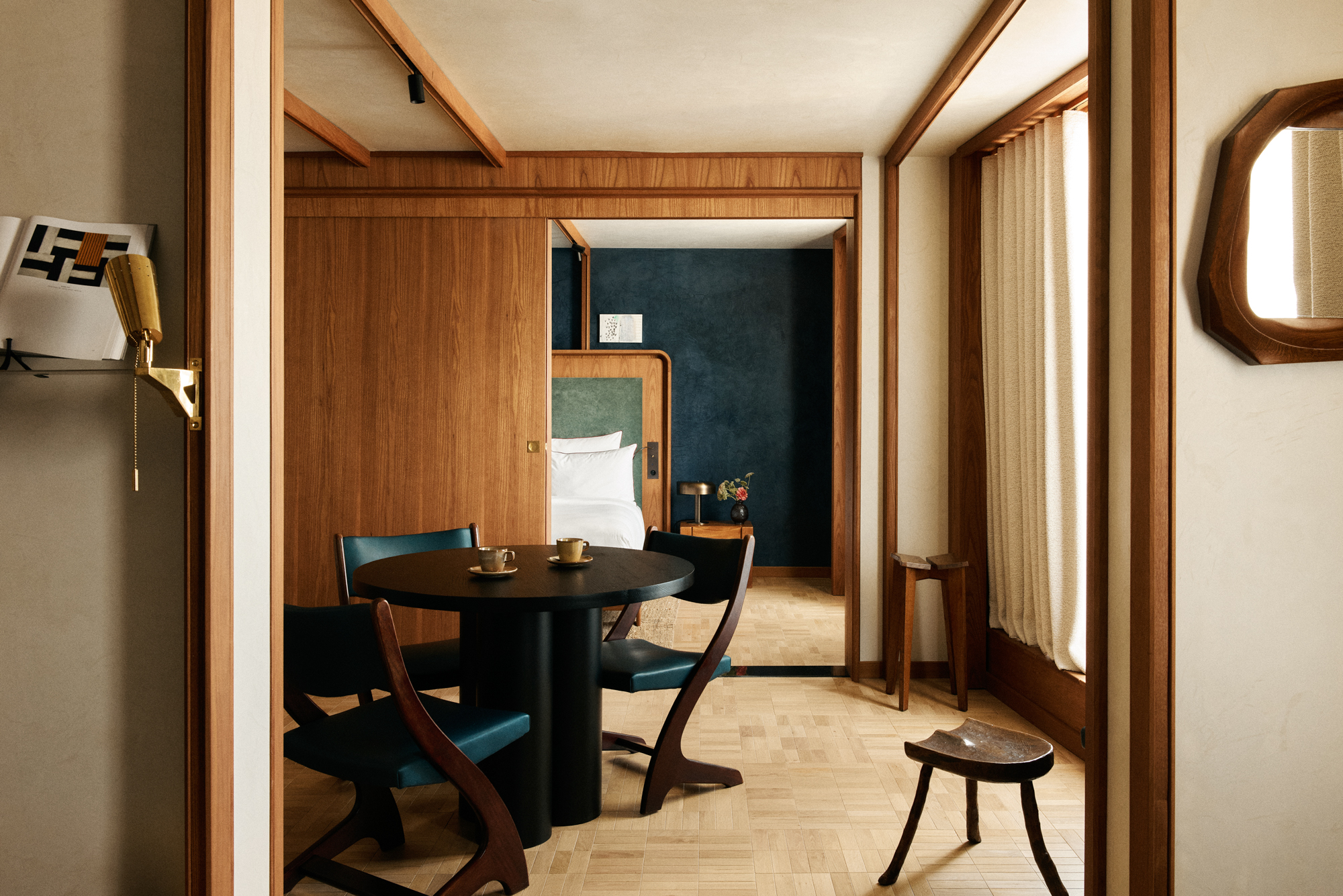 Modern hotel room featuring a round black table with two cups, blue chairs, wooden accents, and a glimpse of a bed in the ...
