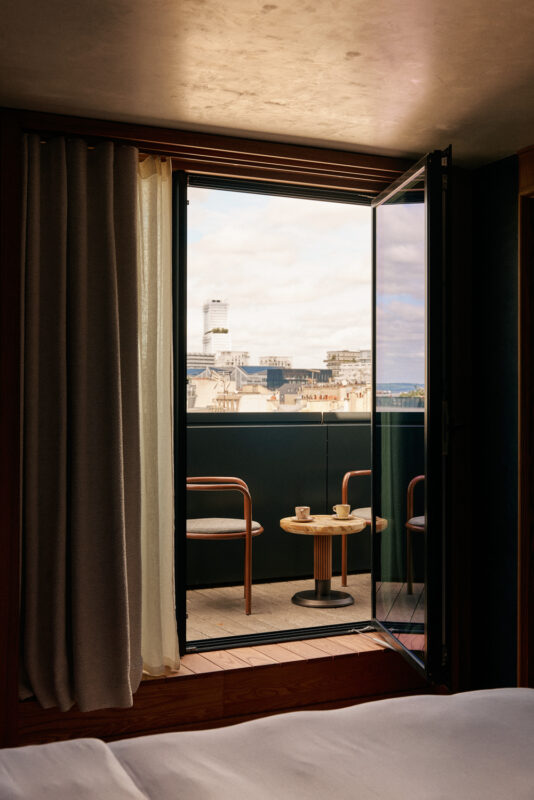 Open balcony door revealing a view of Paris rooftops, with a small table and chairs on the balcony.
