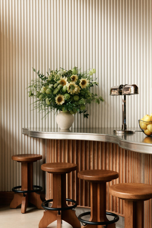 Stylish interior featuring a curved wooden bar, three wooden stools, and a vase of sunflowers against ribbed walls.