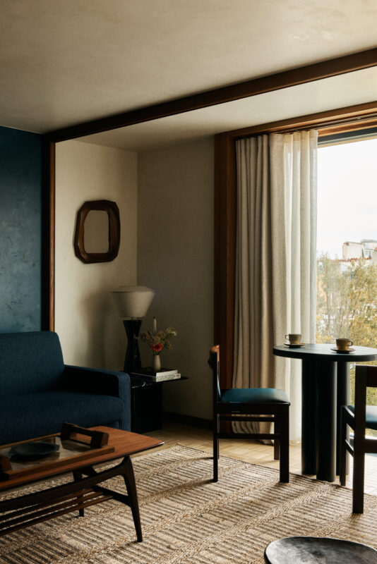 Modern hotel room featuring a blue sofa, wooden table, and a chair, with natural light streaming through large curtains.
